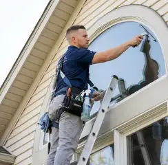 Professional window cleaner on a ladder using a squeegee to clean an exterior arched window, Irvine, CA.